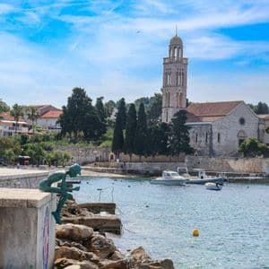 Una statua di bronzo si trova su un molo di cemento che si affaccia su una baia tranquilla con barche e una chiesa in pietra con campanile.
