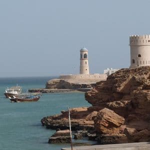 A lighthouse and a watchtower on a rocky coastline overlook a calm bay with two traditional boats.