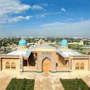 Una vista aerea di un complesso edilizio in mattoni ocra e finemente ornato, caratterizzato da due alti minareti e cupole di un blu brillante, sullo sfondo di un'ampia distesa urbana.