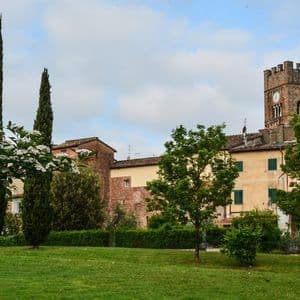 Un parco erboso con cespugli fioriti e cipressi di fronte a vecchi edifici, inclusa una prominente torre dell'orologio in pietra.