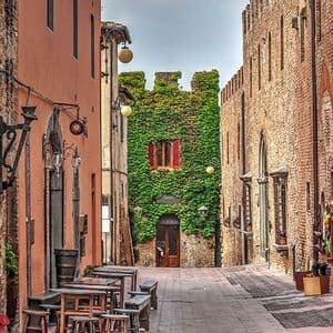 Uno stretto vicolo in mattoni con tavoli da caffè all'aperto conduce a un edificio storico coperto di edera verde.