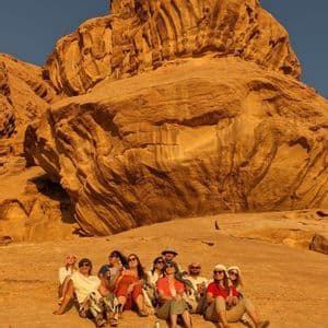 A WeRoad group trip sits together on a vast rock formation in a desert, with a towering cliff behind them.
