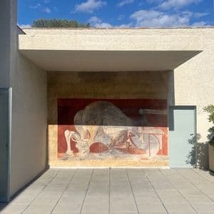 Un murale astratto nei toni del rosso e del grigio sulla parete esterna del patio di un edificio moderno sotto un cielo blu.