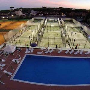 Una vista aerea di un club sportivo al tramonto, con una piscina in primo piano e persone che giocano sui campi da padel illuminati.