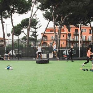 Un gruppo di persone che si allena in una palestra all'aperto su un campo verde in erba sintetica, utilizzando varie attrezzature da fitness come bilancieri e pneumatici.
