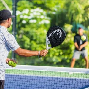 Un uomo visto di spalle serve con una racchetta da padel su un campo all'aperto, con un altro giocatore sullo sfondo.