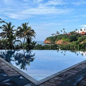 Ein Infinity-Pool mit einem Holzdeck überblickt eine tropische Küste mit Palmen, einer grünen Klippe und dem Ozean.
