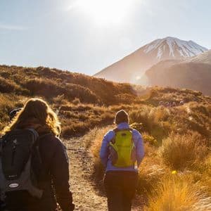 Due escursionisti di un viaggio di gruppo WeRoad camminano su un sentiero soleggiato verso una montagna innevata in lontananza, con altri che li precedono.