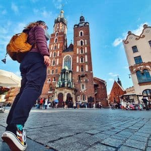Eine Aufnahme aus Froschperspektive einer Person mit gelbem Rucksack, die auf einem Kopfsteinpflasterplatz vor einer großen roten Backsteinkirche mit zwei Türmen steht.