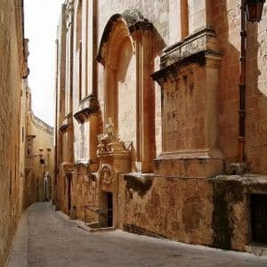 Une ruelle pavée étroite serpente entre de hauts bâtiments historiques en pierre, avec le soleil projetant de fortes ombres.