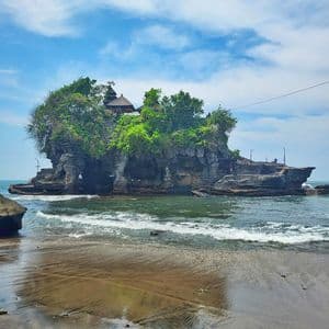 Un templo se asienta sobre un islote rocoso cubierto de árboles en el mar, con suaves olas lamiendo la orilla arenosa.