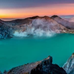 Un lago de cráter volcánico turquesa con vapor elevándose de su caldera rocosa bajo un cielo de amanecer colorido.