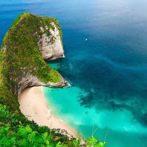 Un gran acantilado cubierto de musgo rodea una recóndita playa de arena blanca con agua turquesa, visto desde un punto elevado.