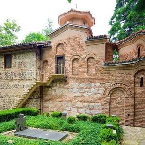 Un edificio storico in mattoni e pietra con una tomba in primo piano, immerso in un rigoglioso giardino verde.