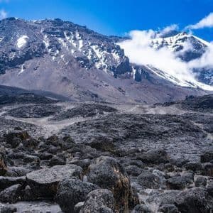 Un vaste paysage rocheux sous un ciel bleu, menant vers une grande montagne avec de la neige et des nuages sur ses sommets.