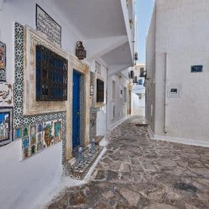 Eine schmale Kopfsteinpflastergasse zwischen weiß getünchten Gebäuden, eines davon mit einer blauen Tür und farbenfrohen, verzierten Keramikfliesen geschmückt.