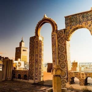 Ornate tiled arches on a rooftop terrace overlook a city skyline with a minaret at sunset.