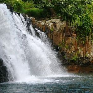 Eine Person schwimmt in einem Pool am Fuße eines breiten Wasserfalls, der über dunkle, säulenartige Felsen fließt und von grünem Laub umgeben ist.