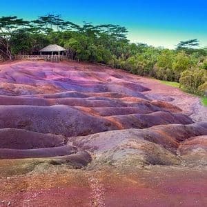 Wellenförmige Dünen aus farbiger Erde mit Lila- und Rottönen, begrenzt von einem grünen Wald und einem hölzernen Aussichtspunkt.