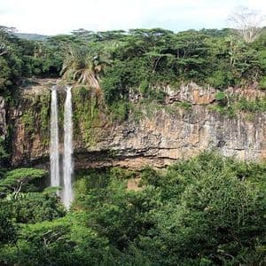 Ein hoher Wasserfall stürzt eine felsige Klippe hinab, umgeben von dichtem, grünem Dschungel unter einem hellen Himmel.