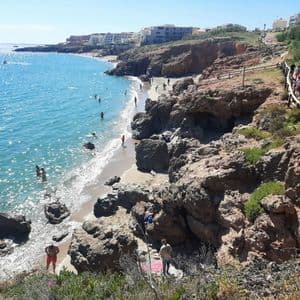Vue plongeante sur une crique rocheuse ensoleillée, avec des personnes nageant dans l'eau bleue et se prélassant sur le sable.