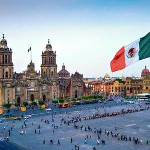 A large Mexican flag waves over a crowded city square in front of a historic cathedral building.