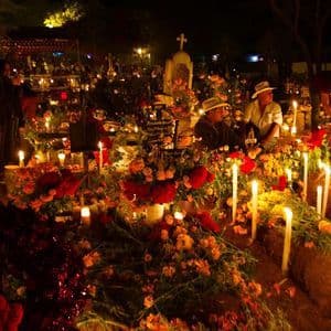 A crowded cemetery at night where graves are decorated with abundant flowers and illuminated by many tall, lit candles.