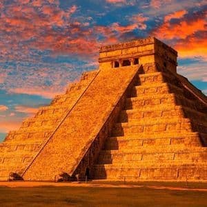 A large stone step-pyramid is illuminated by the setting sun, set against a blue sky with dramatic orange clouds.