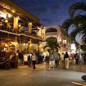 People walk along a busy cobblestone street at dusk, past illuminated multi-story restaurants with outdoor seating and palm trees.
