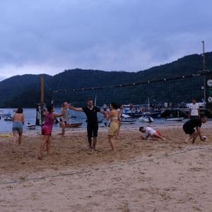 A WeRoad group trip plays beach volleyball on a sandy shore with boats in the water and hills in the background.