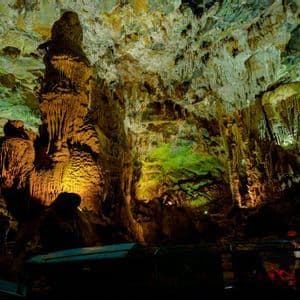 Una barca con un gruppo WeRoad naviga attraverso un'immensa caverna, con stalattiti e stalagmiti illuminate da luci verdi e gialle.