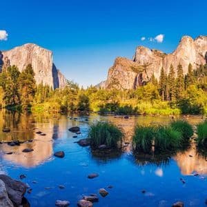 Un fiume calmo con rive rocciose riflette imponenti scogliere di granito e foreste lussureggianti sotto un cielo azzurro brillante.