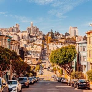 Una ripida strada cittadina con auto parcheggiate e case colorate, che conduce verso un denso skyline sotto un cielo blu.