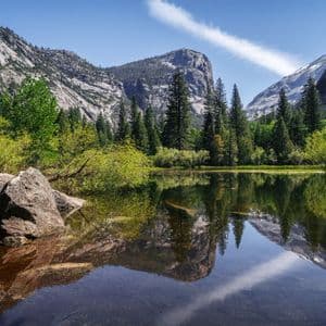 Un lago calmo riflette montagne di granito, una foresta verde e un cielo azzurro in una giornata di sole.