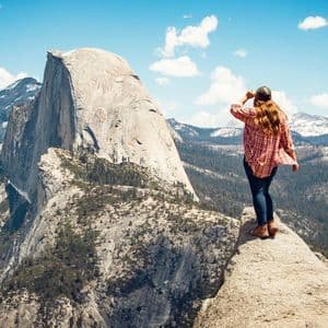 Una donna in camicia a quadri si trova su un punto panoramico roccioso, ammirando una vasta catena montuosa con una cupola di granito sotto un cielo azzurro.