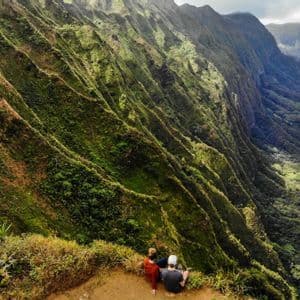 Due persone sono sedute sul bordo di una scogliera, con vista su un vasto paesaggio di montagne ripide dai crinali verdi e una valle sottostante.