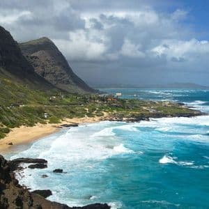 Una costa verde e frastagliata con una spiaggia sabbiosa incontra l'oceano turchese con onde bianche sotto un cielo nuvoloso.
