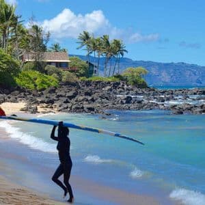 Una persona in muta trasporta una longboard sulla testa lungo la riva di una spiaggia tropicale con palme.