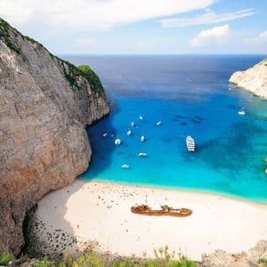 Ein rostiges Schiffswrack liegt an einem weißen Sandstrand in einer Bucht, umgeben von hohen Klippen, mit Booten, die auf türkisem Wasser schwimmen.