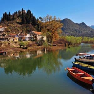 Barche colorate ormeggiate su un lago calmo di fronte a un villaggio collinare con una fortezza sulla cima sotto un cielo sereno.