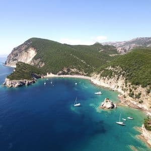 Vista aerea di una baia turchese con barche a vela all'ancora, racchiusa tra ripide scogliere alberate sotto un cielo azzurro e limpido.