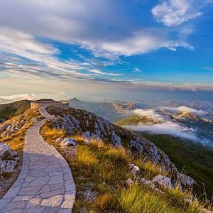 Un sentiero lastricato in pietra si snoda lungo un crinale roccioso di montagna verso un punto panoramico circolare, che si affaccia su una vasta valle con nuvole al di sotto.