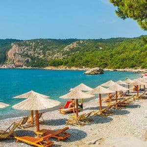 File di ombrelloni di paglia e lettini in legno costeggiano una spiaggia di ciottoli accanto all'acqua turchese, con una collina boscosa sullo sfondo.