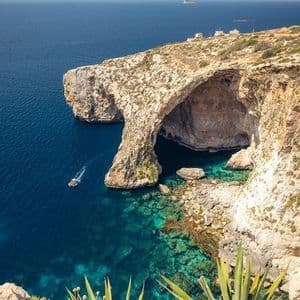 Vista dall'alto di una costa rocciosa con un grande arco naturale sull'acqua turchese, dove una piccola barca naviga.