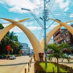 Ein großes Denkmal aus gekreuzten Elefantenstoßzähnen wölbt sich über einer Stadtstraße mit Autos, Palmen und Gebäuden unter blauem Himmel.