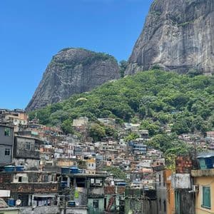 Un'ampia veduta di edifici colorati di una favela, ammassati su una verde collina di fronte a ripide montagne rocciose, sotto un cielo azzurro.