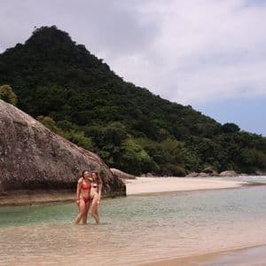 Dos mujeres de un viaje en grupo de WeRoad sonríen en la orilla de una playa tropical con una colina boscosa al fondo.