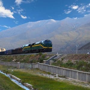 Un treno merci verde e giallo viaggia su un binario attraverso una vasta valle di montagna sotto un cielo blu con nuvole bianche.