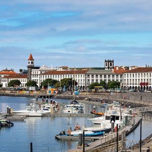 Une marina remplie de bateaux amarrés le long d'un quai en pierre, avec une ville côtière de bâtiments blancs et une tour de l'horloge en arrière-plan.