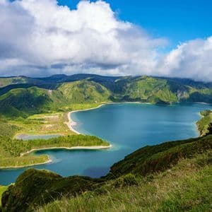 Vue plongeante sur un grand lac bleu au cœur d'un cratère volcanique, entouré de pentes montagneuses verdoyantes, sous un ciel bleu parsemé de nuages blancs.